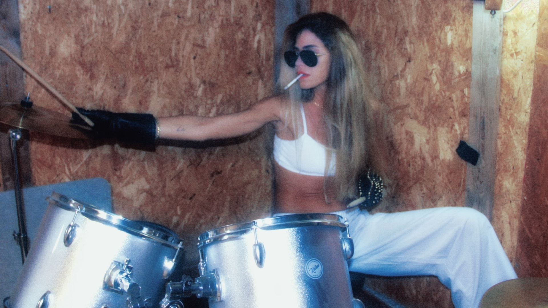 Woman wearing black studded leather gloves and white pants sits at a drum set, legs apart, poised and powerful in a dimly lit room.