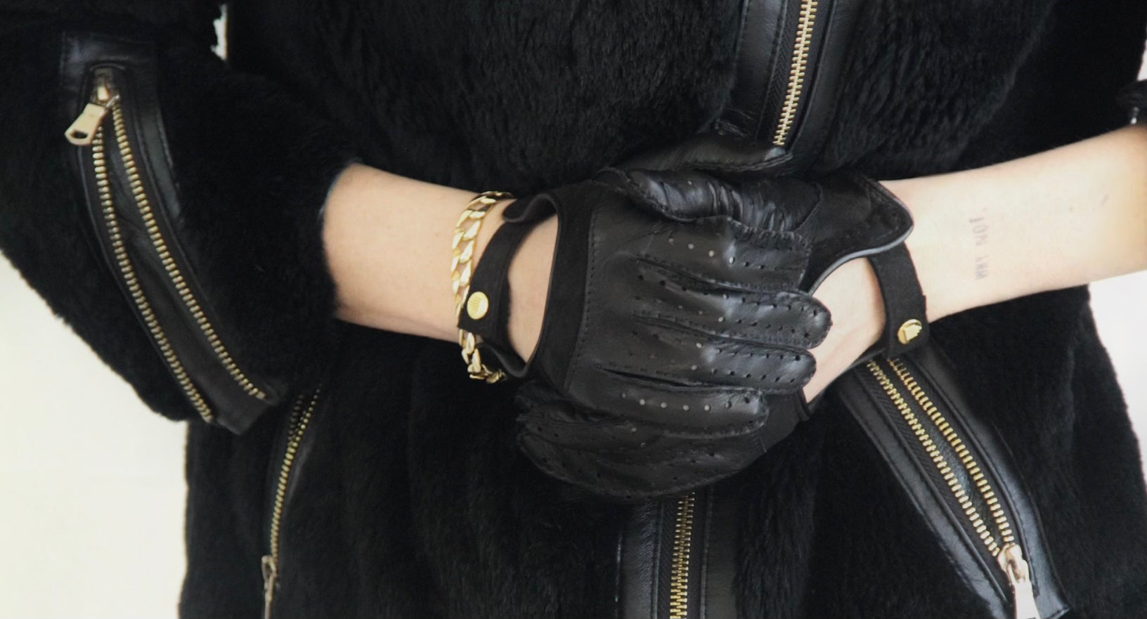 Close-up of a woman’s hands wearing black leather driving gloves with cutout knuckles, layered with a gold chain bracelet, against a black textured coat with gold zippers.
